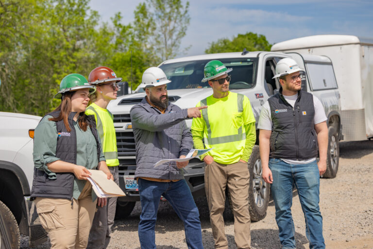 Five construction workers in hard hats and safety vests gathered by trucks, one man pointing while others hold plans.