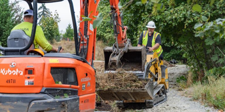 Construction workers in high-visibility vests operating orange excavator and small loader clearing vegetation on a gravel path