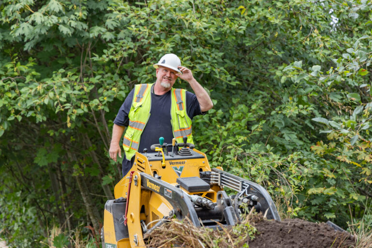 Construction worker in a hard hat and high-visibility vest standing behind a yellow Vermeer machine in leafy woods