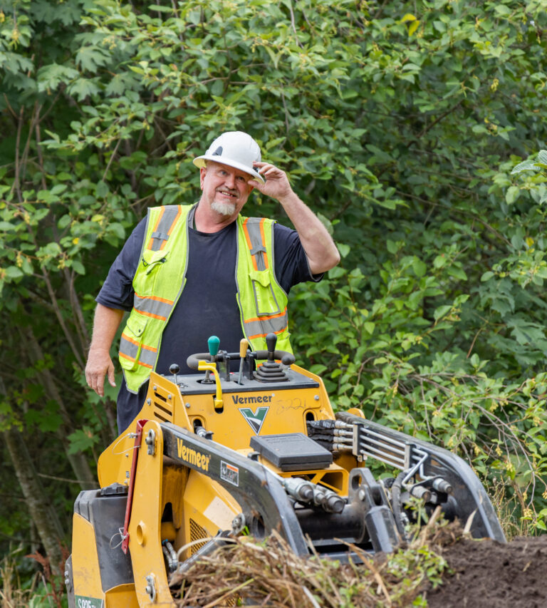 Construction worker in a hard hat and high-visibility vest standing behind a yellow Vermeer machine in leafy woods