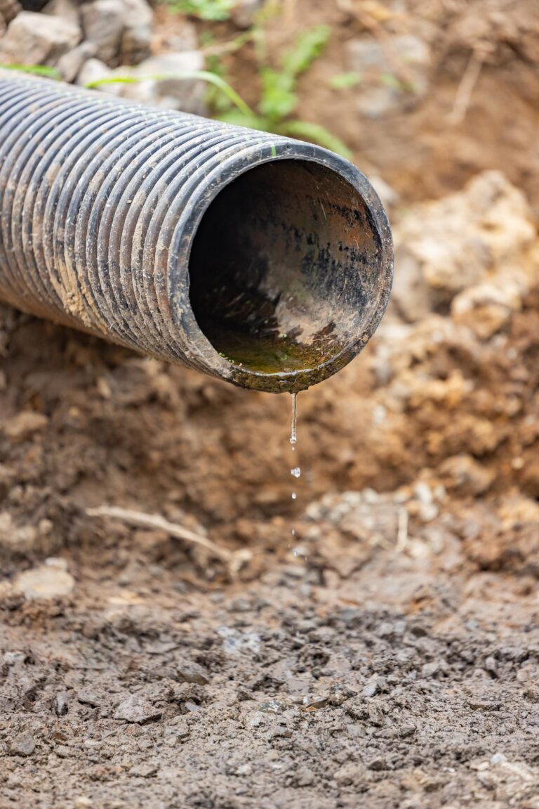 Corrugated black drainage pipe tipped over on dirt with a few drops of water falling from its open end.
