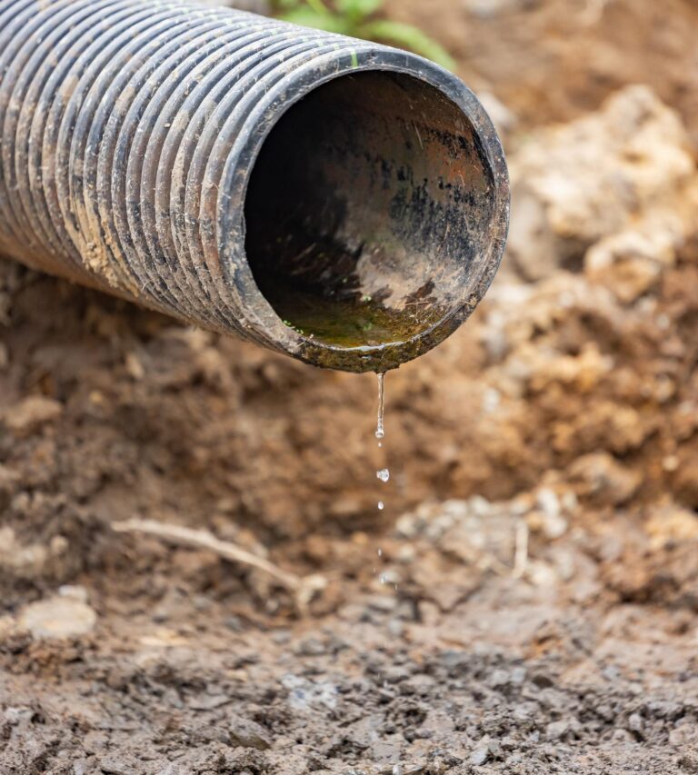 Corrugated black drainage pipe tipped over on dirt with a few drops of water falling from its open end.