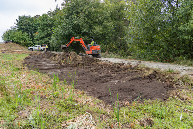 Orange mini excavator digging a narrow dirt path at edge of a wooded area while workers and trucks stand nearby