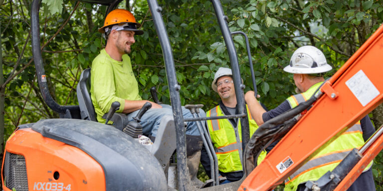 Three construction workers in hard hats and high-visibility vests chatting around an orange mini excavator in a leafy setting.