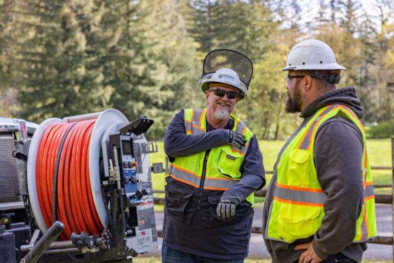 Two construction workers in hard hats and high-visibility vests talking beside a truck-mounted orange hose reel in a park-like setting.