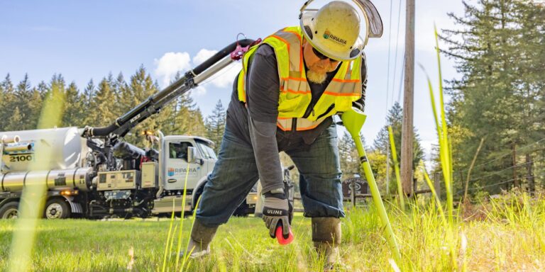 Worker in a safety vest and hard hat using a tool in grassy roadside with utility truck and trees in background