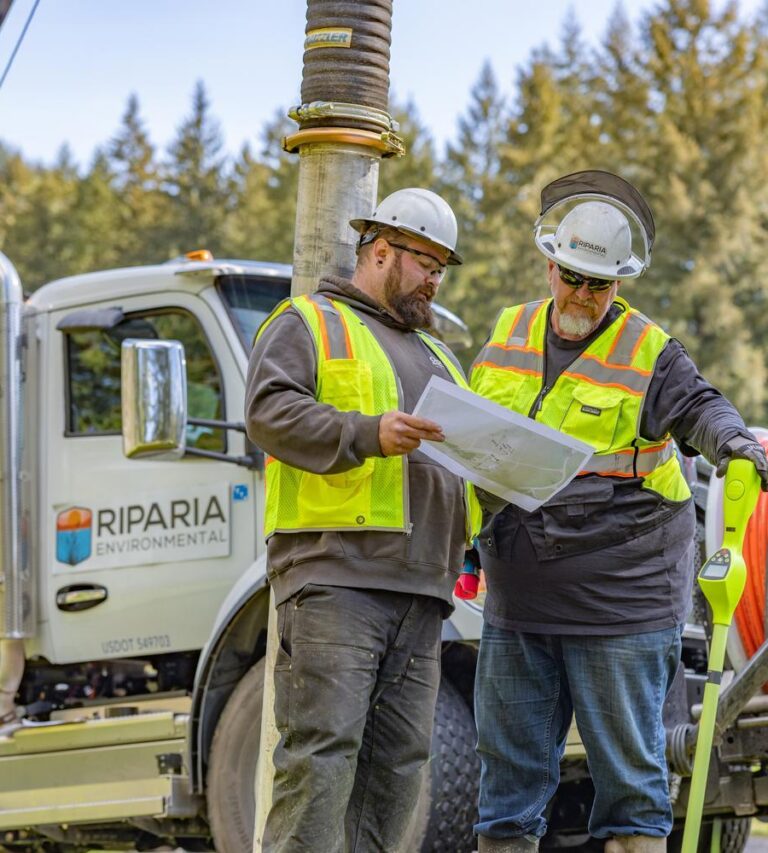 Two construction workers in hard hats and hi-vis vests review plans beside a Riparia Environmental service truck with equipment.