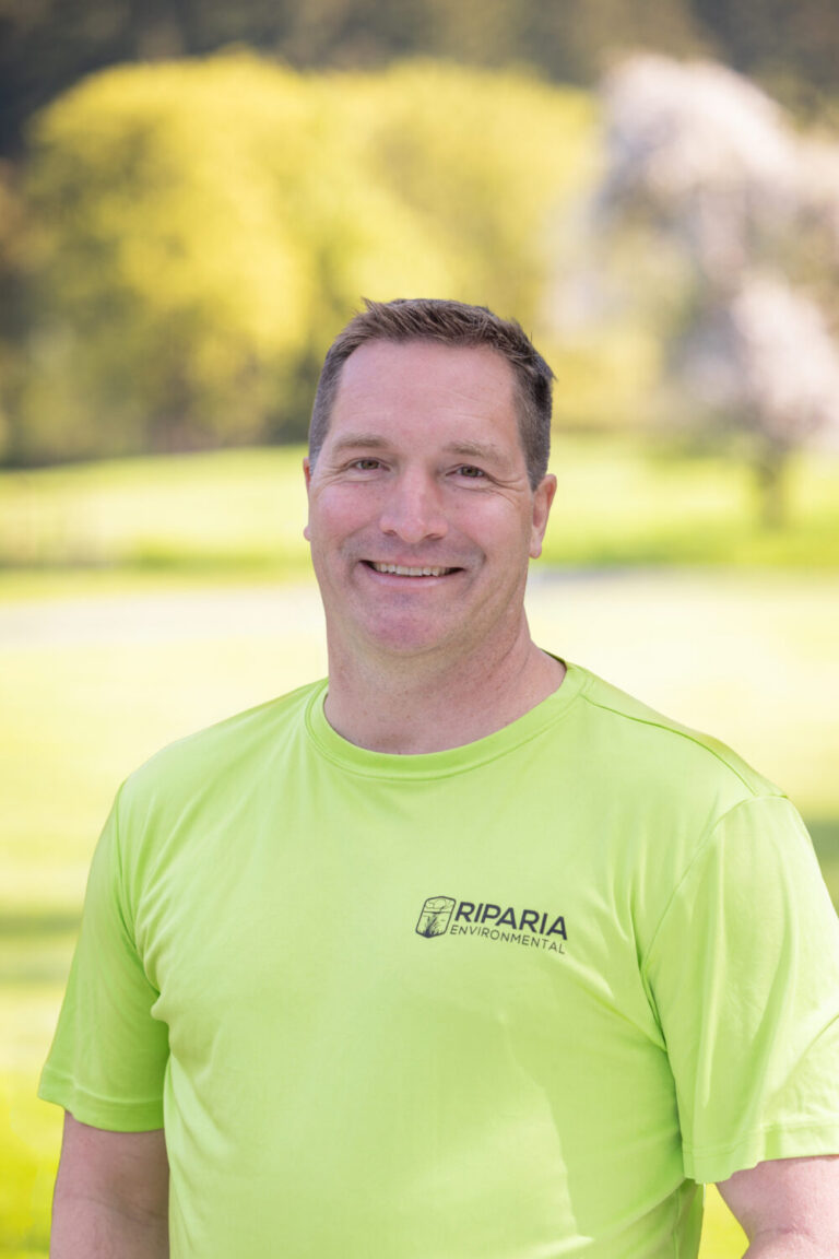 Smiling middle-aged man wearing a bright lime green Riparia Environmental t-shirt, standing outdoors with blurred trees and grass behind him