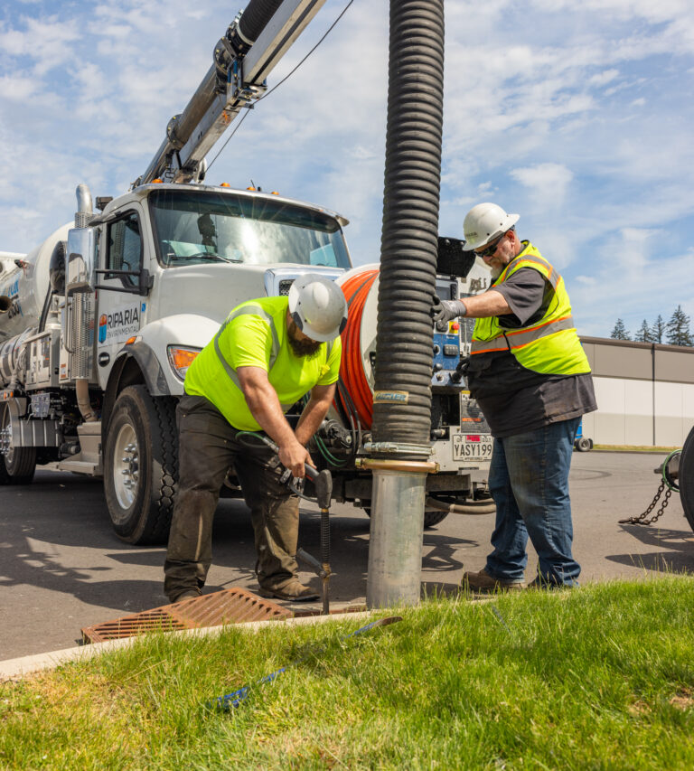 Two utility workers in high-visibility vests and helmets operate a large vacuum truck hose while opening a storm drain on a sunny day.