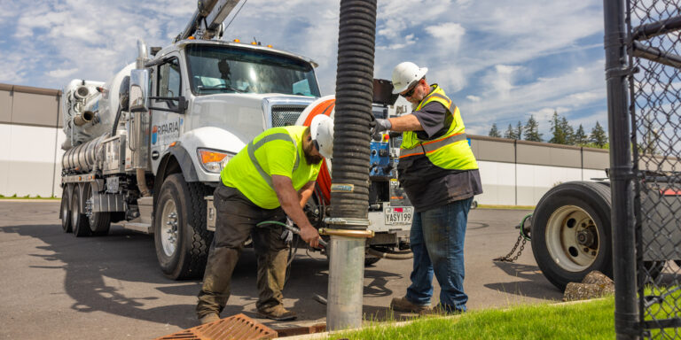 Two utility workers in hard hats and high-visibility vests operating a large vacuum truck hose over a storm drain in a parking lot.