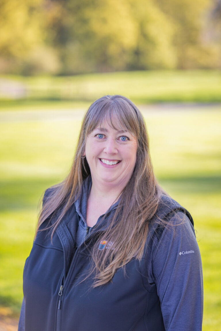 Smiling middle-aged woman with long light brown hair and blue eyes wearing a dark Columbia jacket, standing outdoors on a sunny grassy background