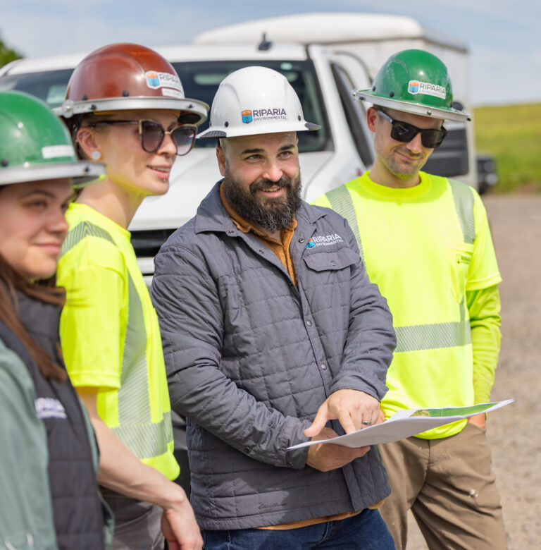 Four construction workers wearing hard hats and high-visibility shirts review plans while standing beside a white work truck.