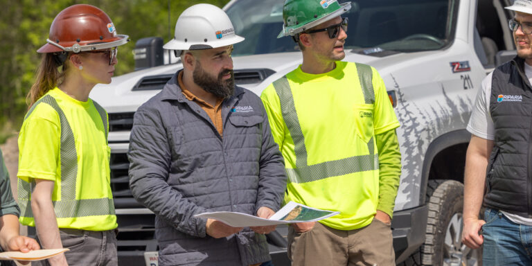 Four construction workers in hard hats and high-visibility shirts standing by a white pickup, one holding project plans.