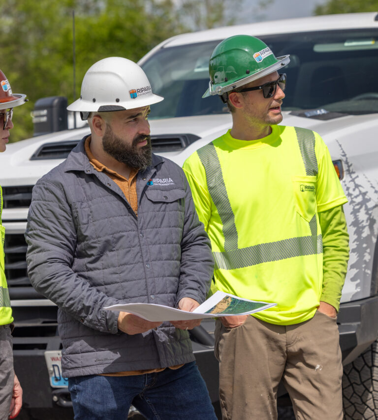 Four construction workers in hard hats and high-visibility shirts standing by a white pickup, one holding project plans.