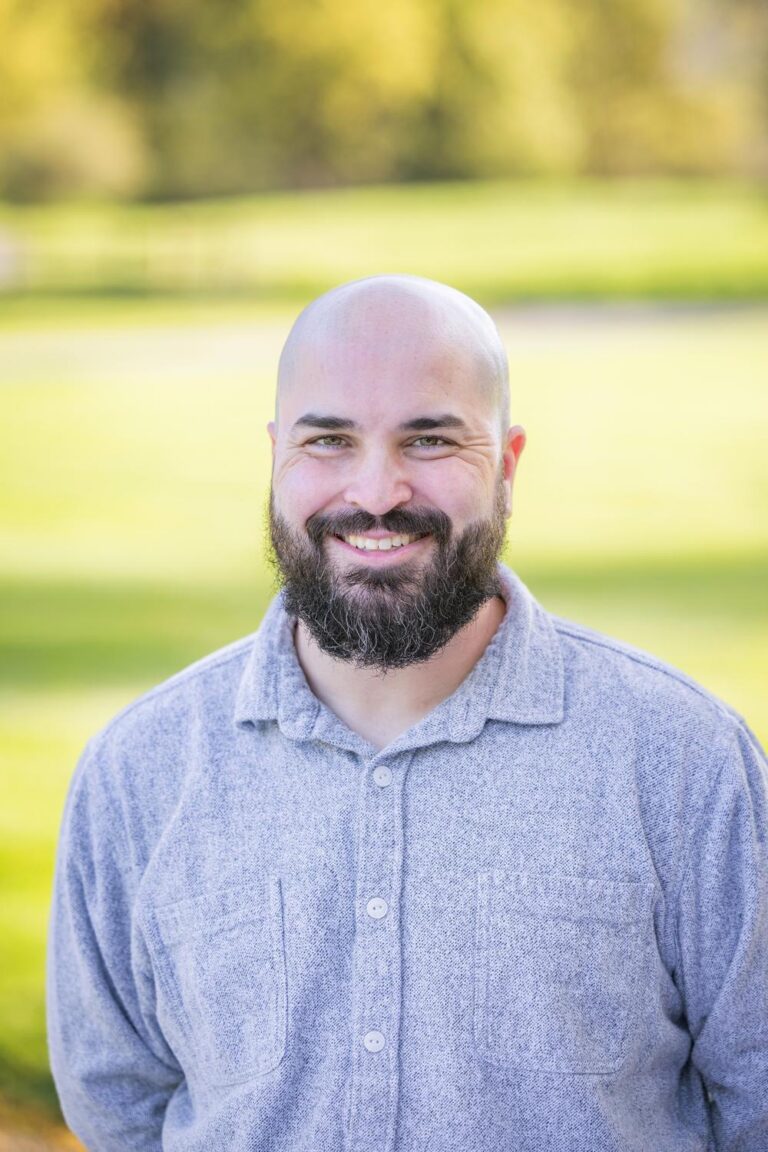 Smiling bald man with a full dark beard wearing a light gray button-up shirt, standing outdoors with blurred green background